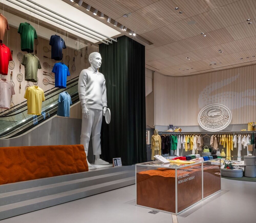 Ground floor of Lacoste Flagship Store, Fifth Avenue, New York, with clothing displays and a large founder statue holding a tennis racket, illuminated by Zeplinn’s architectural lighting.